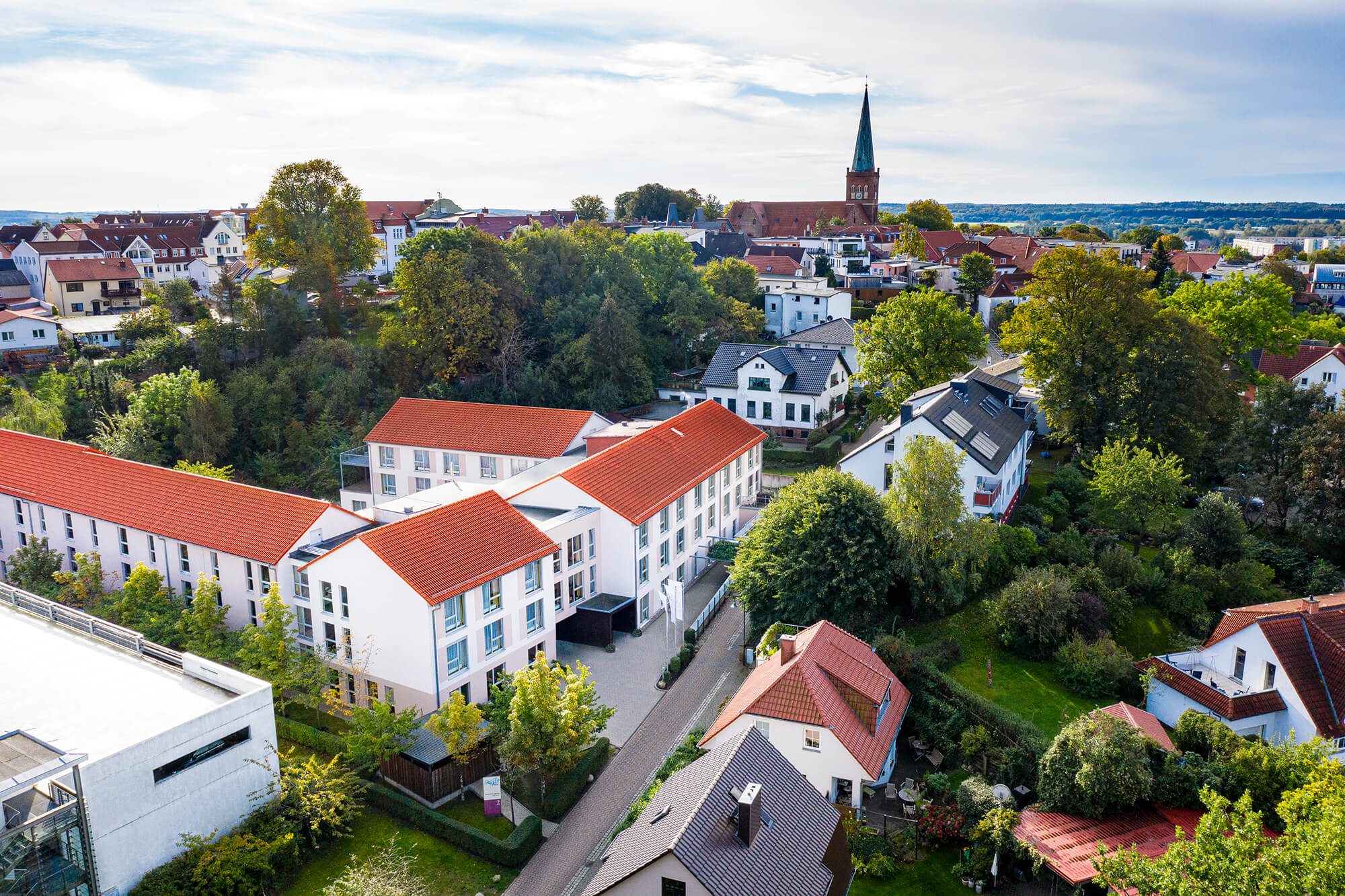 Luftaufnahme eines modernen Gebäudekomplexes mit roten Dächern in einer ruhigen Wohngegend, umgeben von Bäumen und kleineren Häusern.