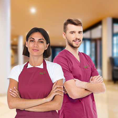 Eine Frau und ein Mann in bordeauxfarbener Berufskleidung stehen mit verschränkten Armen nebeneinander in einem modernen, hellen Innenraum. Beide blicken selbstbewusst in die Kamera – die Szene vermittelt Professionalität und Teamgeist im Pflege- oder Se