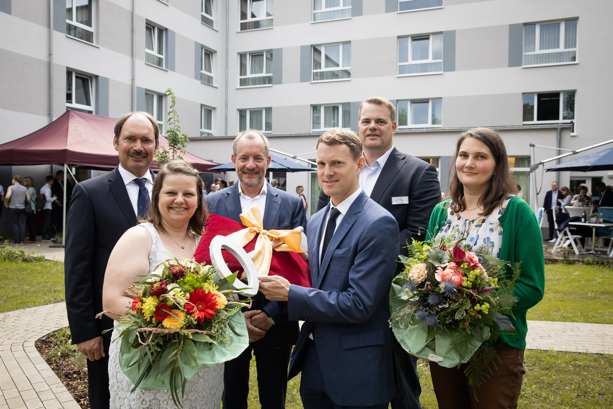 Gruppenfoto von sieben festlich gekleideten Personen bei einer feierlichen Veranstaltung im Innenhof eines modernen Gebäudekomplexes. Drei Frauen halten Blumensträuße, ein Mann überreicht symbolisch einen großen Schlüssel. Die Stimmung wirkt freudig und f